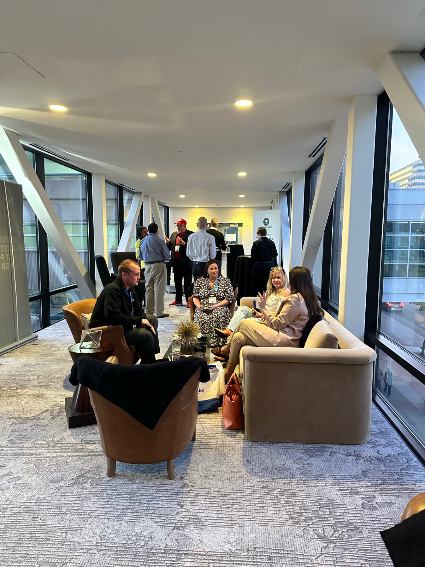 Guests seated in lounge chairs and standing in small conversation groups along the glass-walled Hotel Ivy skyway